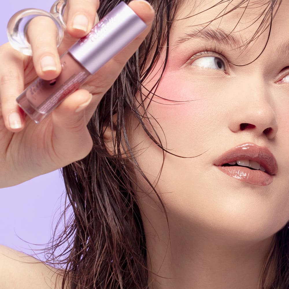 Close-up of a woman holding a mauve lip-gloss tube near her face, glossy lips, pink blush and wet hair against a lilac background