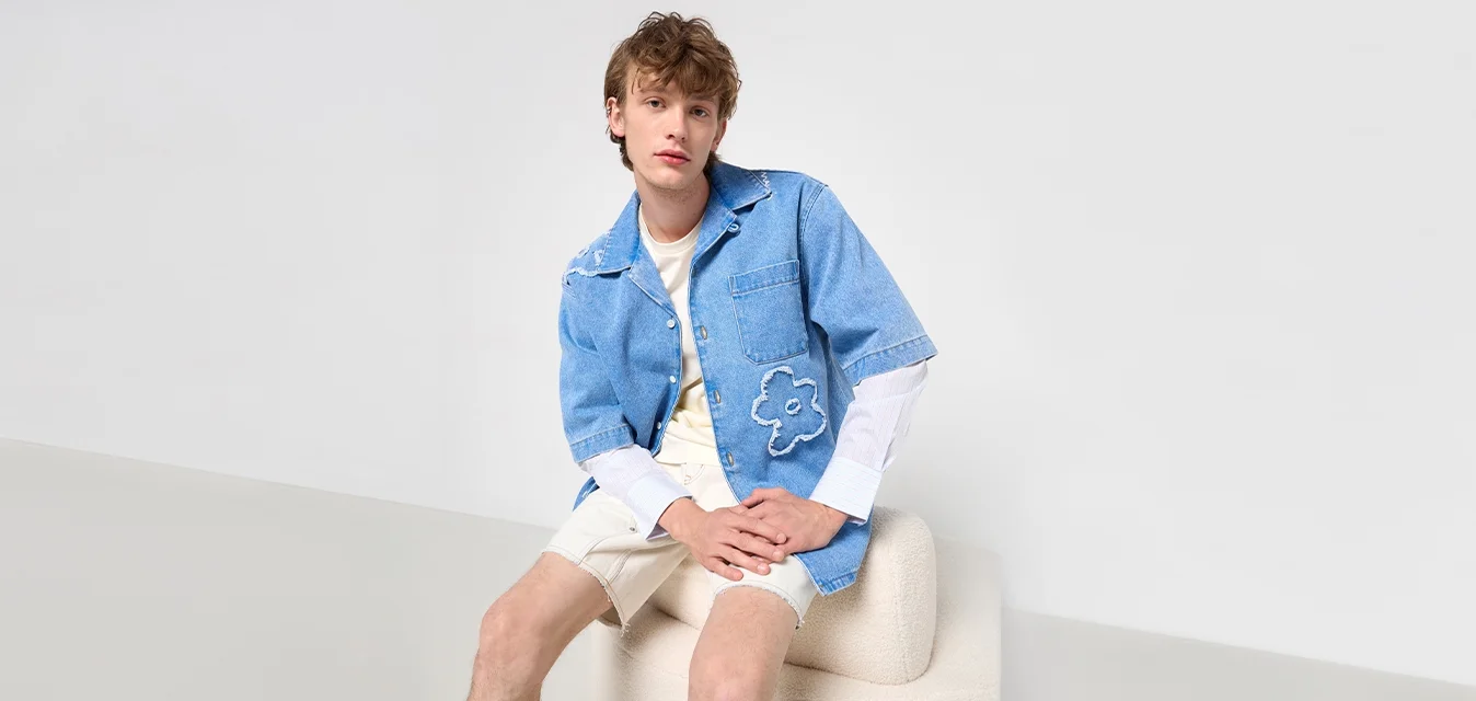 Camicia corta in denim blu con taschino sul davanti e ricamo floreale, sovrapposta a una maglietta bianca a maniche lunghe; abbinata a pantaloni corti beige.