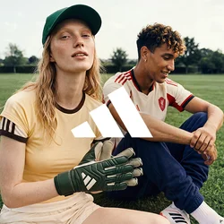 Una donna che indossa guanti da portiere e un cappellino è seduta sull’erba accanto a un uomo in abbigliamento sportivo su un campo aperto con alberi e edifici sullo sfondo.