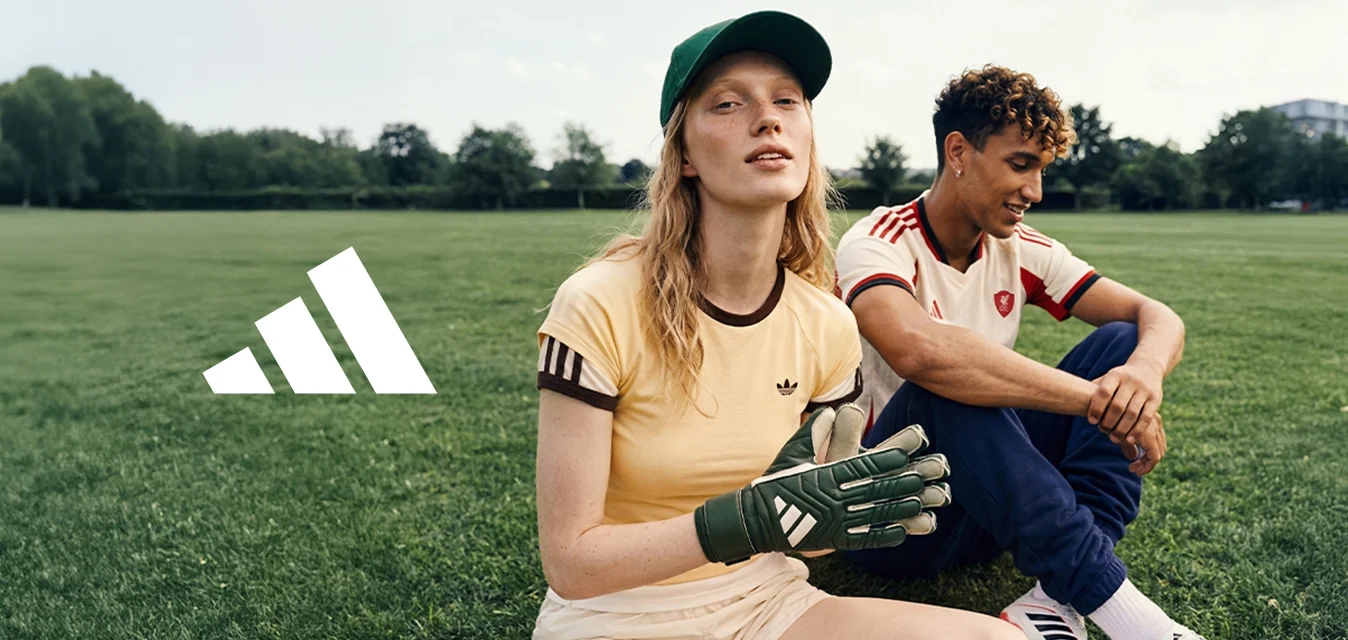 Una donna che indossa guanti da portiere e un cappellino è seduta sull’erba accanto a un uomo in abbigliamento sportivo su un campo aperto con alberi e edifici sullo sfondo.