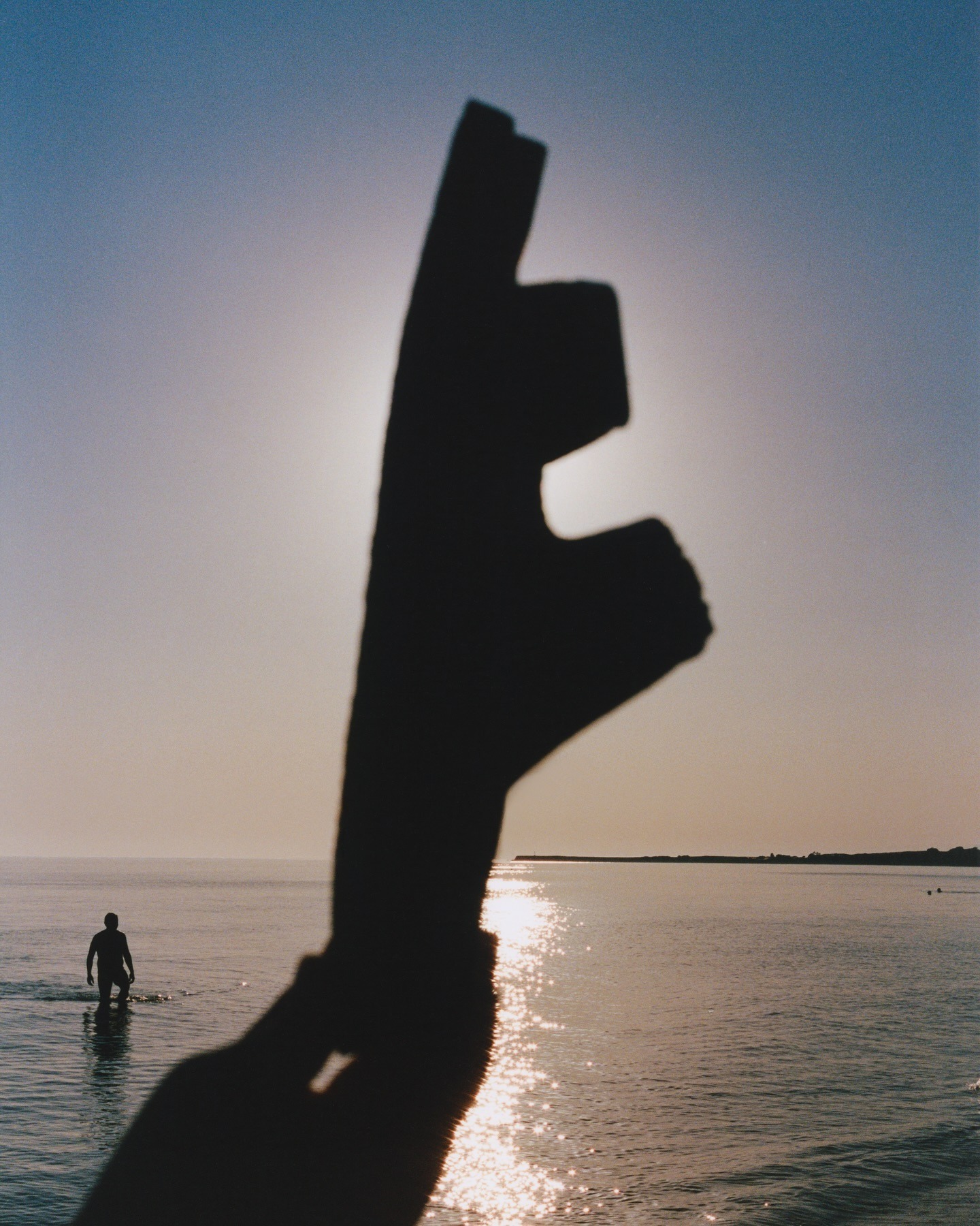 Silhouette of hand-shaped driftwood held against the sun over a shimmering sea, with a person wading in the distance