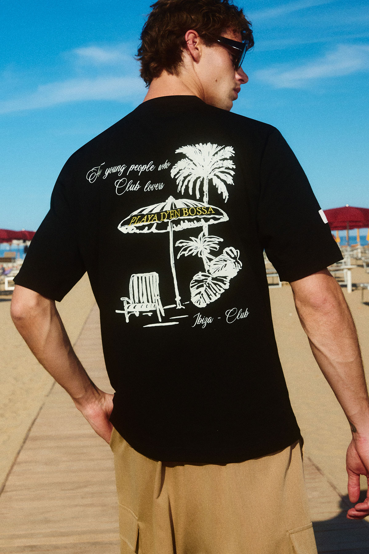 Young man wearing black t-shirt with palm-tree and umbrella print, standing on wooden boardwalk at a sunny beach, wearing sunglasses