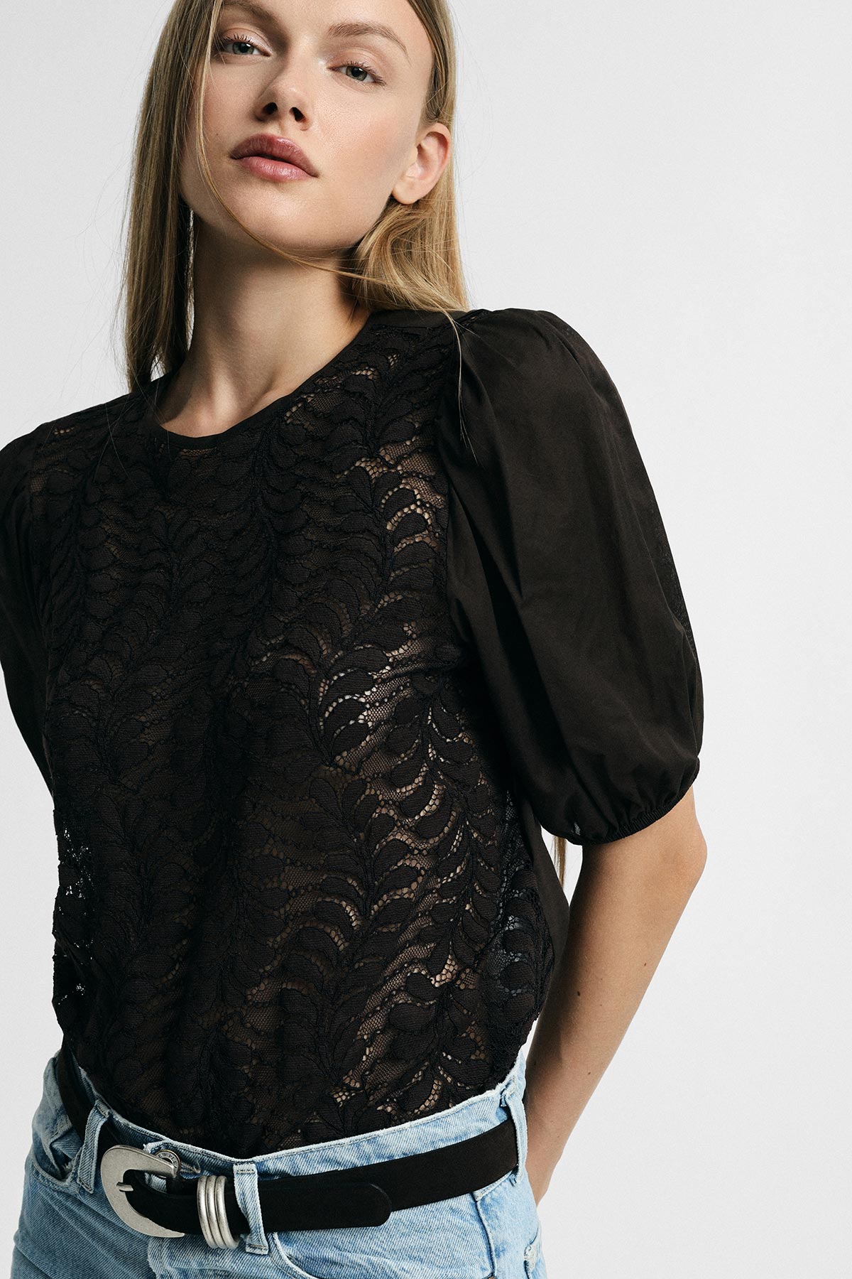 Young woman modelling black lace blouse with puff sleeves, tucked into light denim jeans with black belt against a neutral background