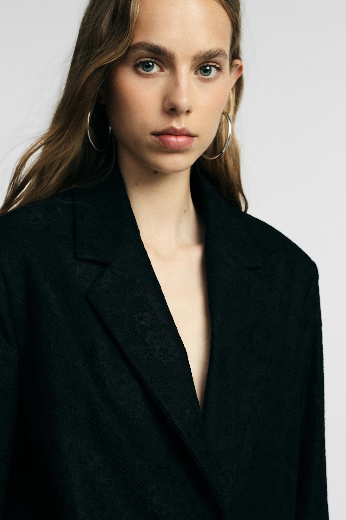 Close-up portrait of a young woman model wearing a black blazer and large hoop earrings against a neutral background, minimalist fashion look