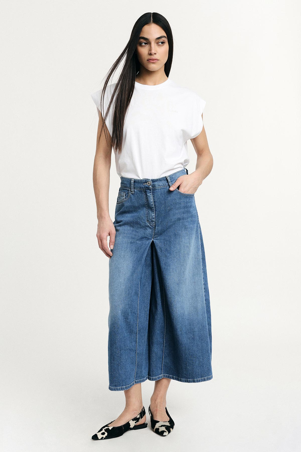 Woman in a white sleeveless T‑shirt tucked into wide‑leg cropped denim culottes and black‑white patterned flats, standing against plain background