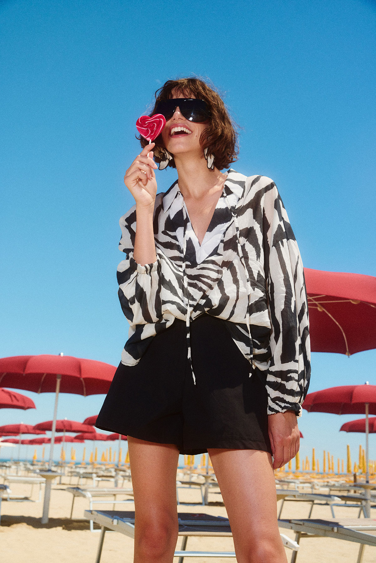 Smiling woman in sunglasses and zebra-print blouse holding a heart-shaped lollipop on a sunny beach with rows of red umbrellas