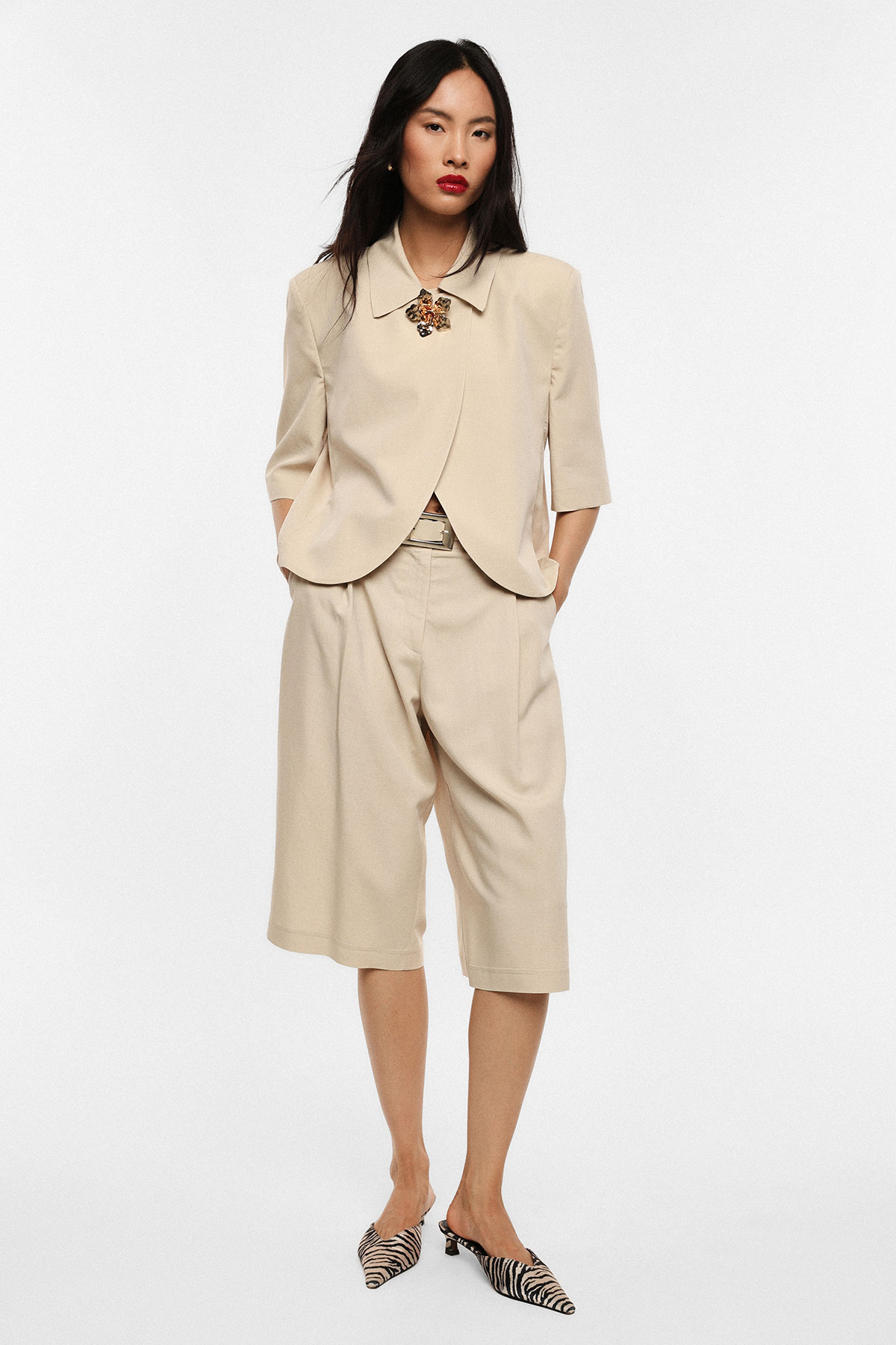 Woman wearing beige short-sleeve jacket and wide bermuda shorts, zebra-print mules and statement brooch, hands in pockets