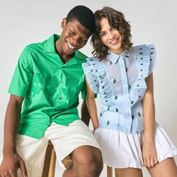 Giovane uomo in camicia verde e pantaloni beige e giovane donna in blusa blu con volant e gonna bianca siedono vicini, sorridendo davanti a uno sfondo semplice.