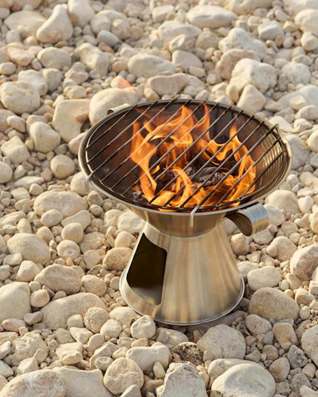 Portable stainless-steel charcoal barbecue with visible flames on a small metal grill, sitting on a rocky pebble beach
