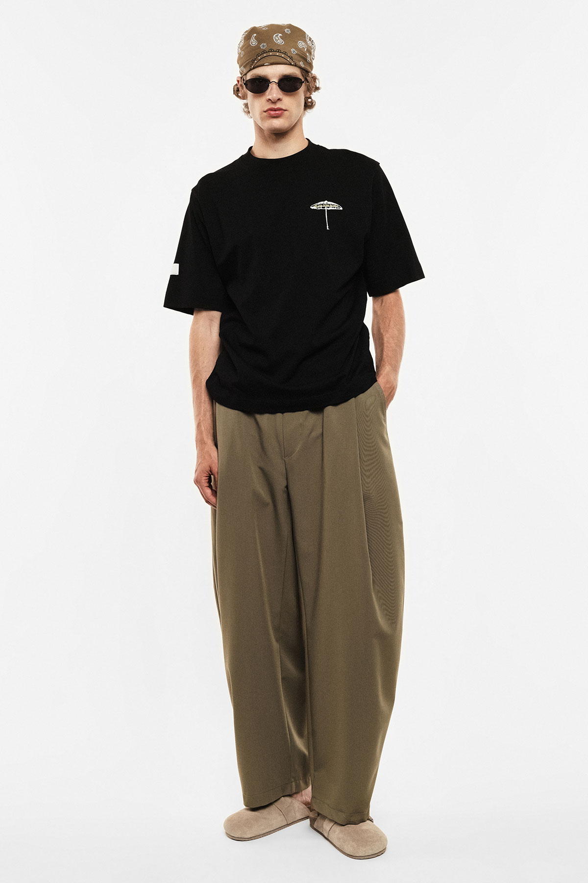 Male model in black t-shirt with small umbrella logo, oversized khaki trousers, bandana and sunglasses on plain white background