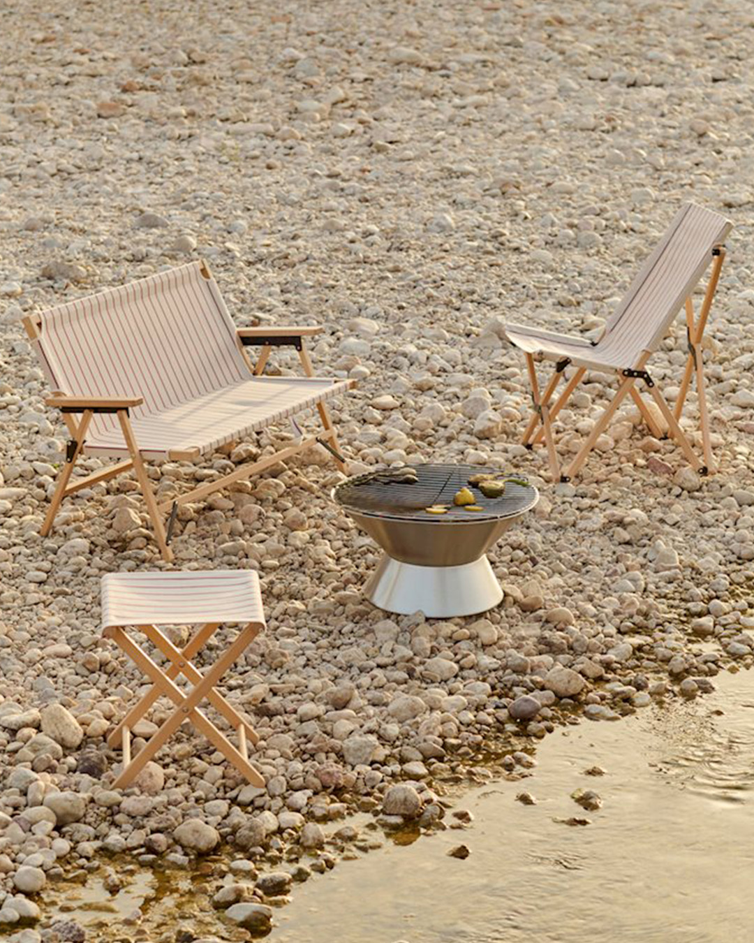 Pebble beach scene with wooden folding chairs, small stool and a portable firepit grill beside the water