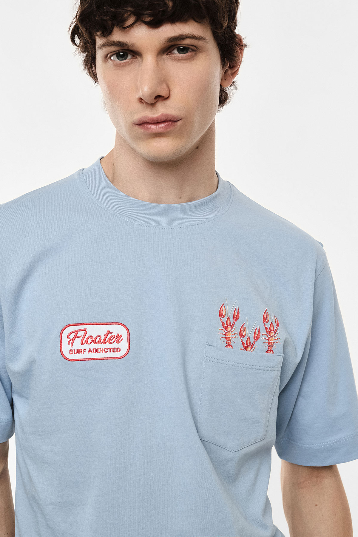 Young man wearing a light blue T‑shirt with a chest pocket, red lobster embroidery and 'Floater Surf Addicted' patch