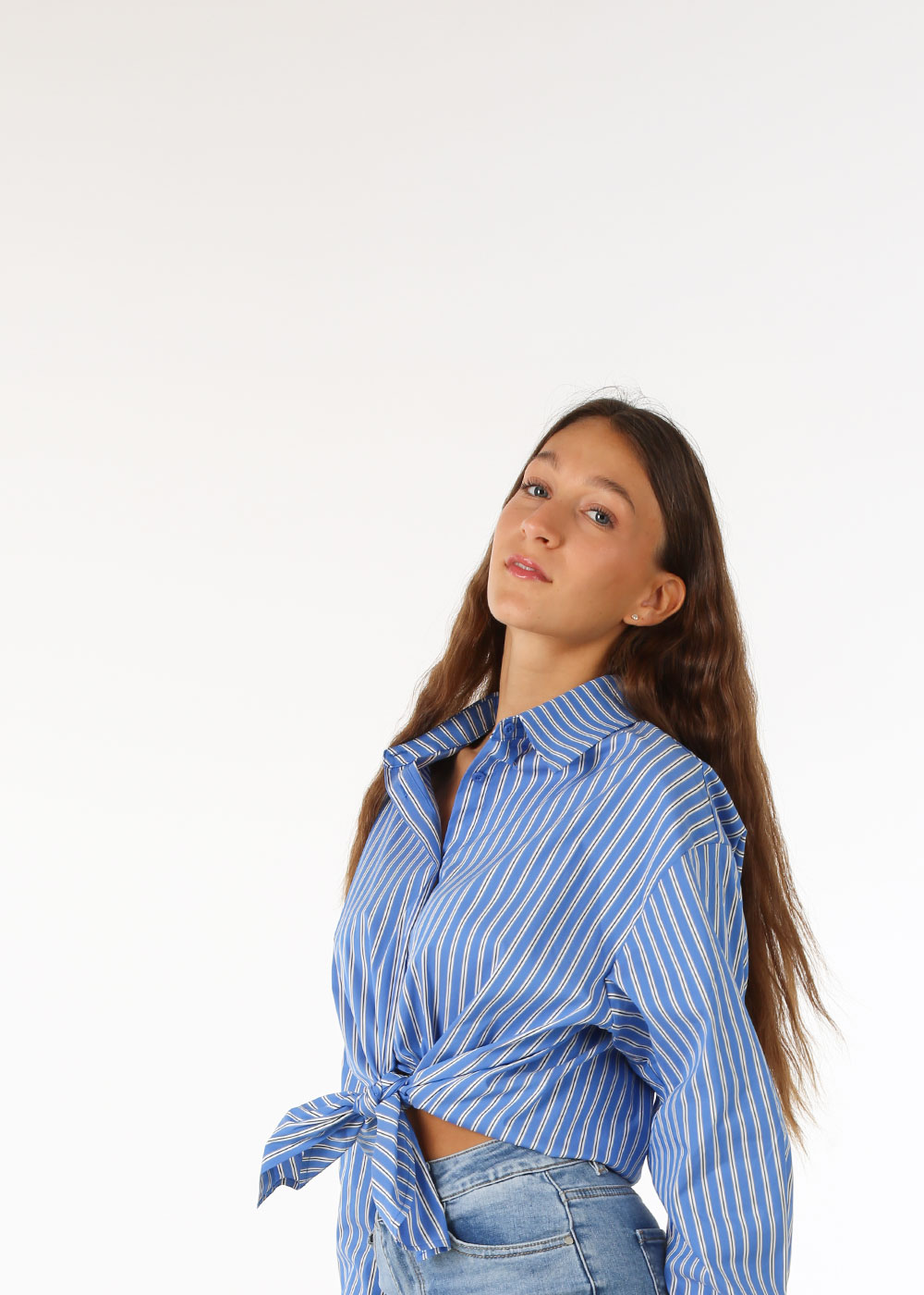 Young woman wearing a blue striped tie-front shirt and jeans posing against a plain white background