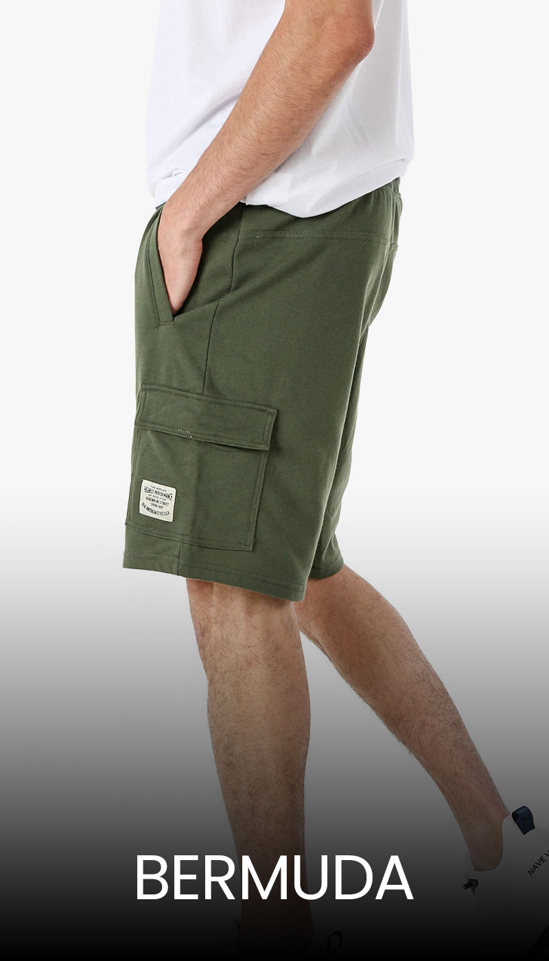 Man wearing olive green cargo Bermuda shorts with side flap pocket and white T‑shirt, hand in pocket and white trainers visible