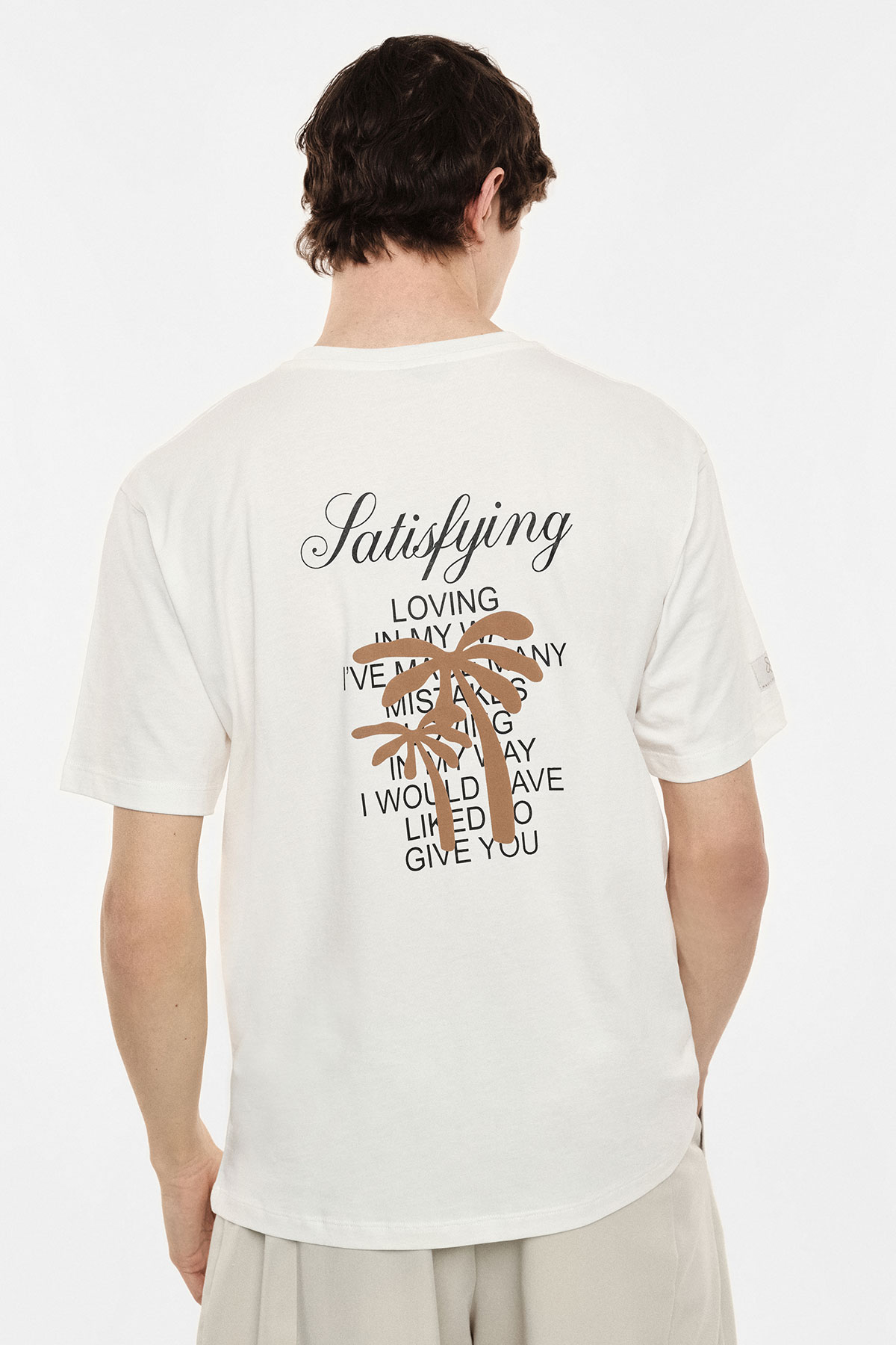 Person seen from back wearing a white t-shirt with the word "Satisfying" in script, a brown palm graphic and stacked black text printed on the back