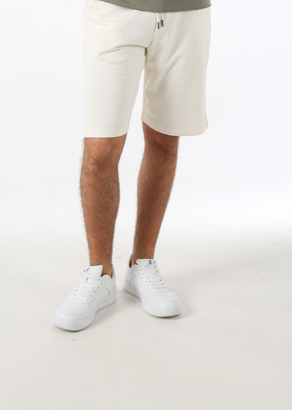 Men's off-white mid-length drawstring cotton shorts worn with white low-top trainers on model against plain background