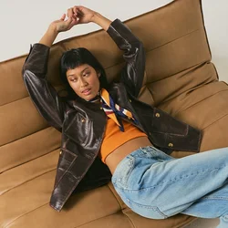 Donna sdraiata su una grande poltrona imbottita marrone da terra, che indossa una giacca nera, una maglia arancione, jeans blu e stivali con tacchi alti color camoscio.