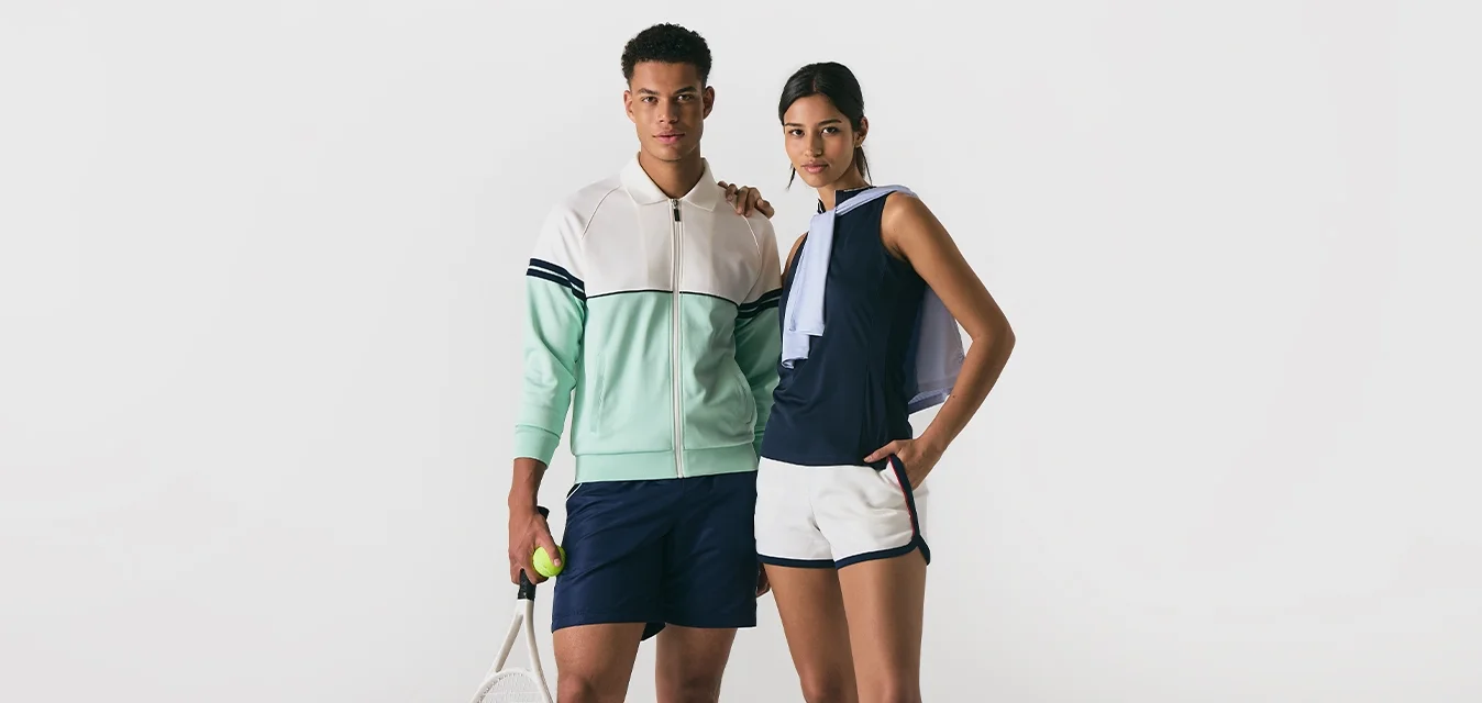 Giovane uomo e donna in abbigliamento da tennis in piedi uno accanto all'altro, l'uomo tiene una racchetta e una palla, la donna ha una mano in tasca e una giacca sulle spalle.