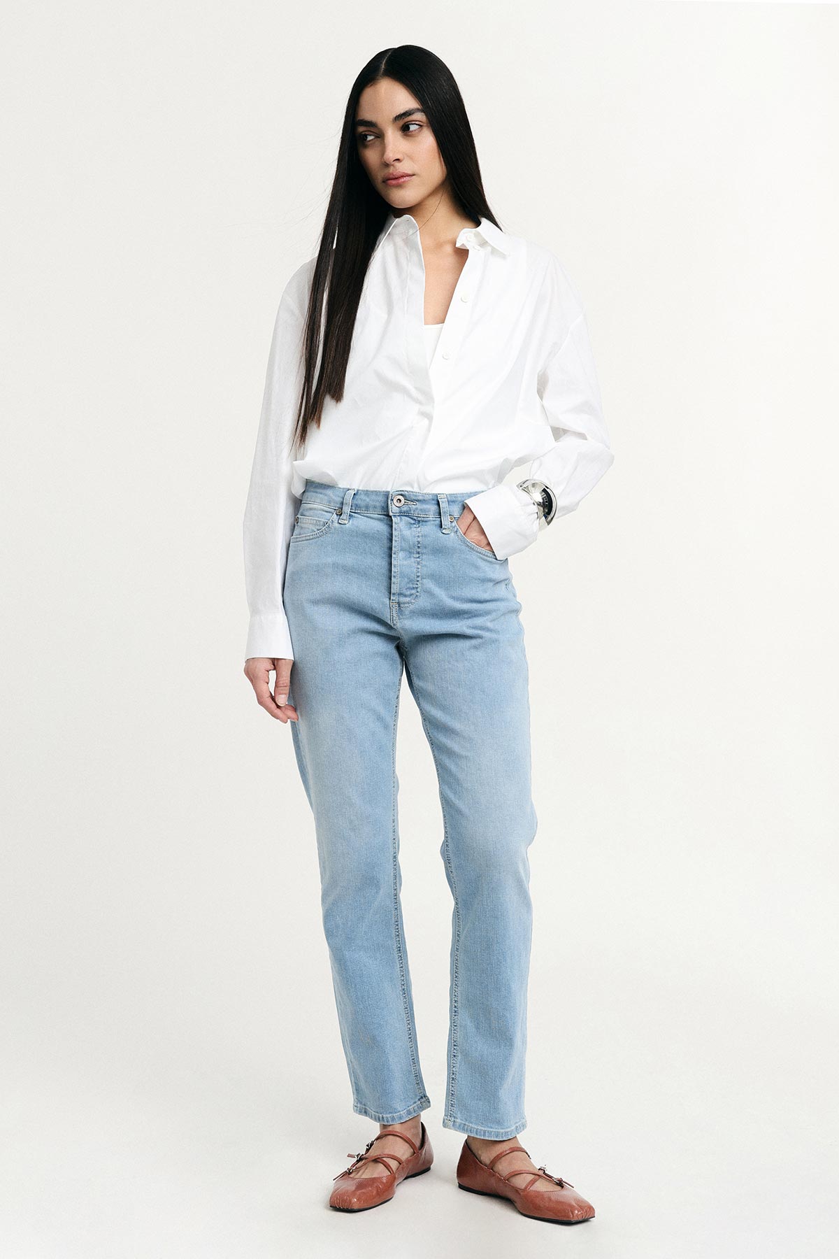 Woman with long dark hair wearing a white shirt, light-blue straight-leg jeans and brown leather flats against a plain white background