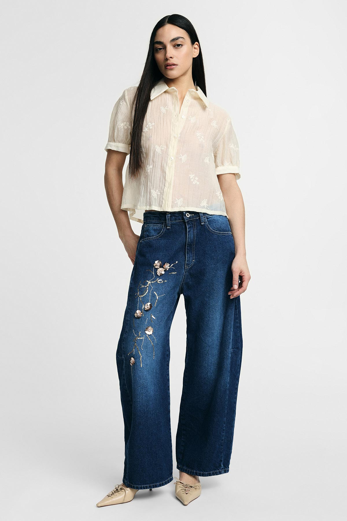 Young woman wearing a sheer cream short-sleeve blouse and wide-leg dark blue jeans with floral embroidery and beige pointed shoes