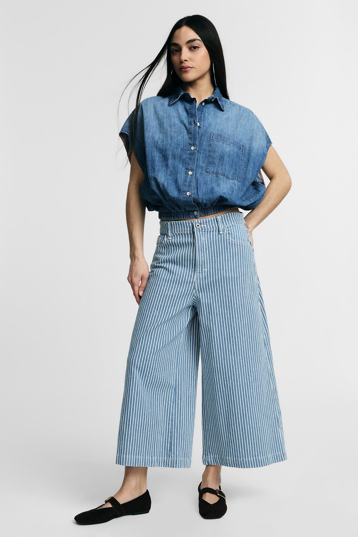 Woman wearing a denim short-sleeve shirt tucked into high-waisted blue-and-white striped wide-leg culottes and black flats, standing