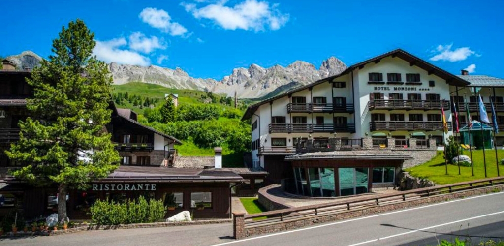 Alpine hotel and ristorante by a roadside with balconies, green slopes and rugged mountains under a bright blue sky