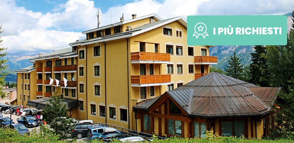 Yellow alpine hotel with wooden balconies and octagonal glass pavilion, parked cars and pine trees against mountain backdrop