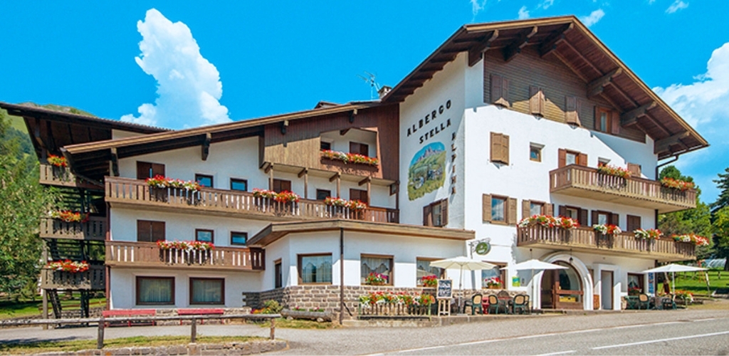 Albergo Stella Alpine hotel with wooden balconies, colourful flower boxes and outdoor seating under a bright blue sky