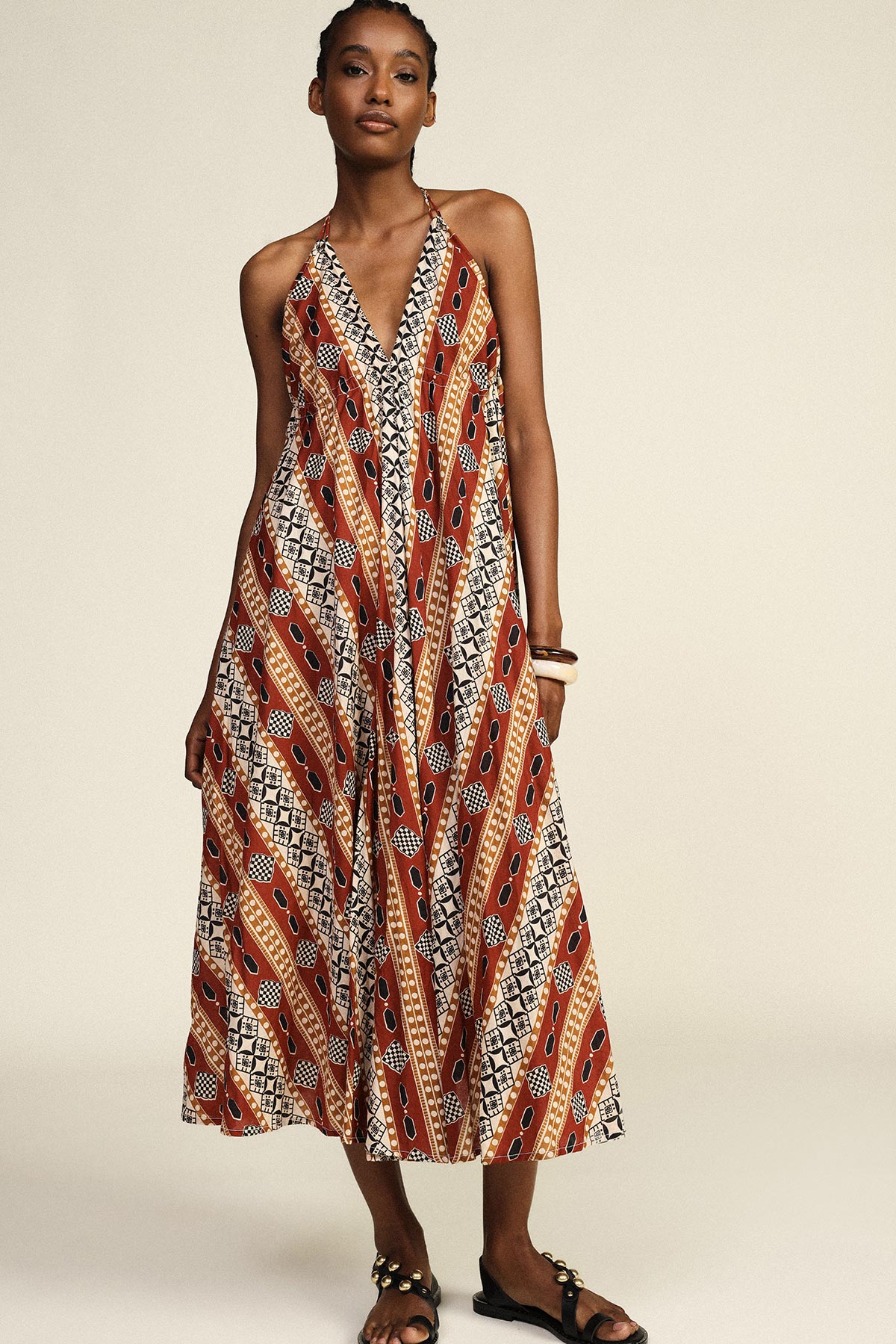 Model wearing a sleeveless halter midi dress with rust, black and cream geometric print and sandals