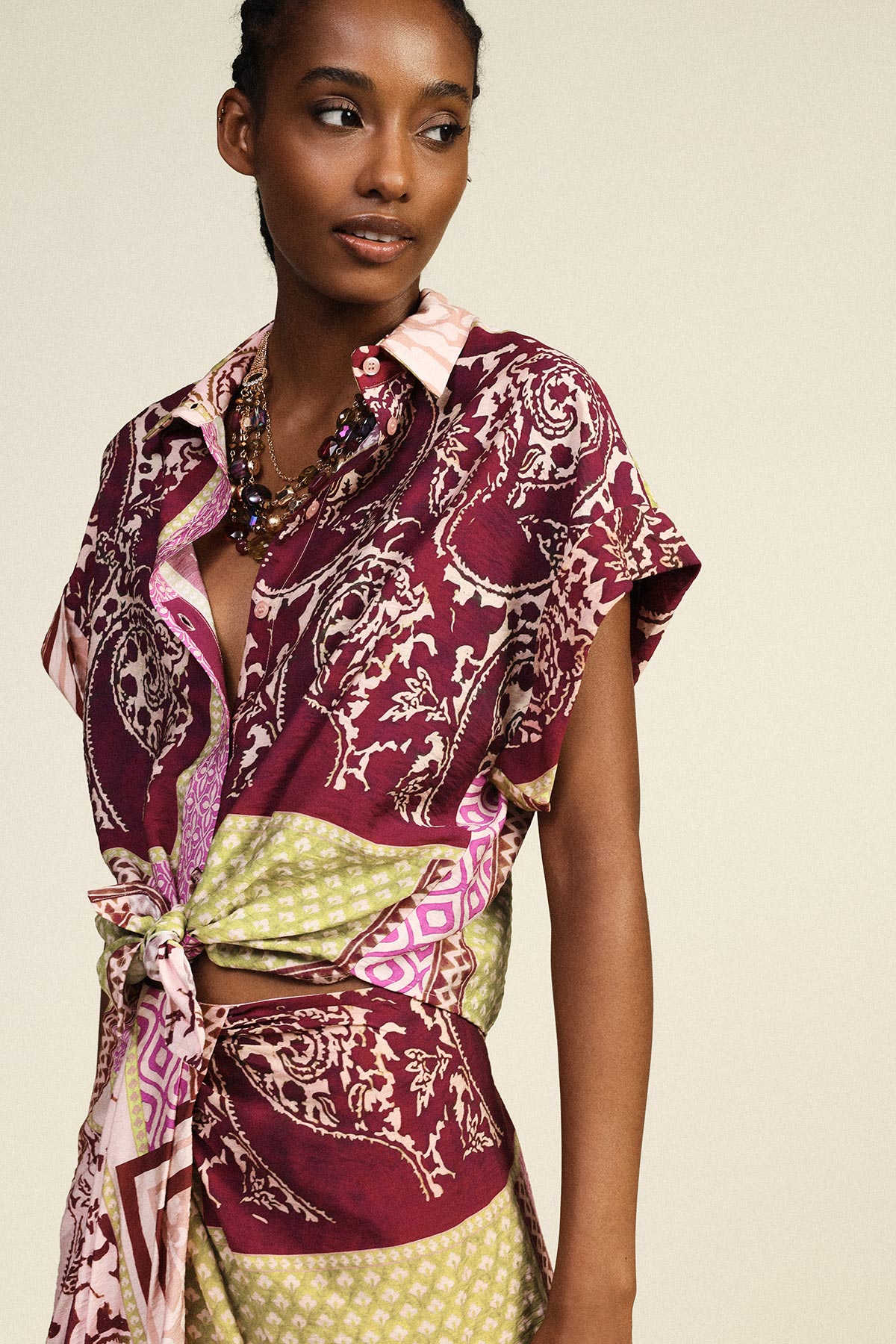 Woman modelling burgundy, pink and green patterned tie-front blouse and matching skirt with beaded necklace, looking to the side