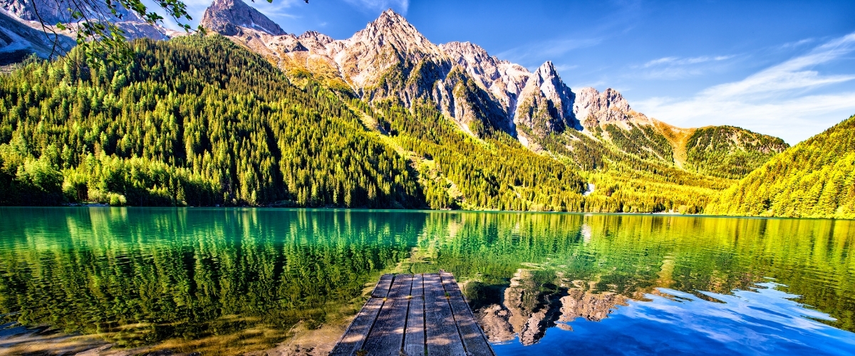 Wooden jetty leading to a turquoise alpine lake with evergreen forest and rugged mountains reflected beneath a clear blue sky