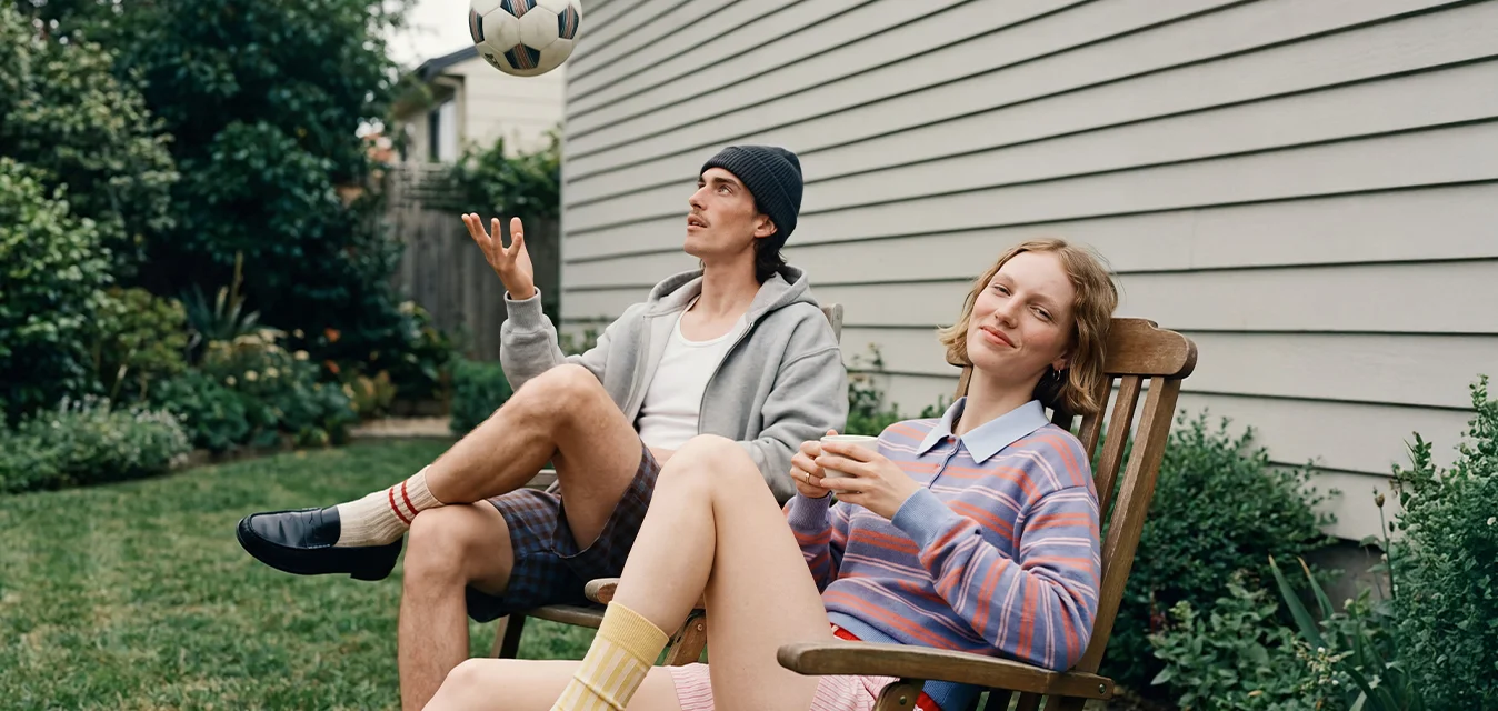 Un uomo fa giocoleria con un pallone da calcio mentre è seduto sull'erba, e una donna si rilassa su una sedia a sdraio di legno in un giardino.