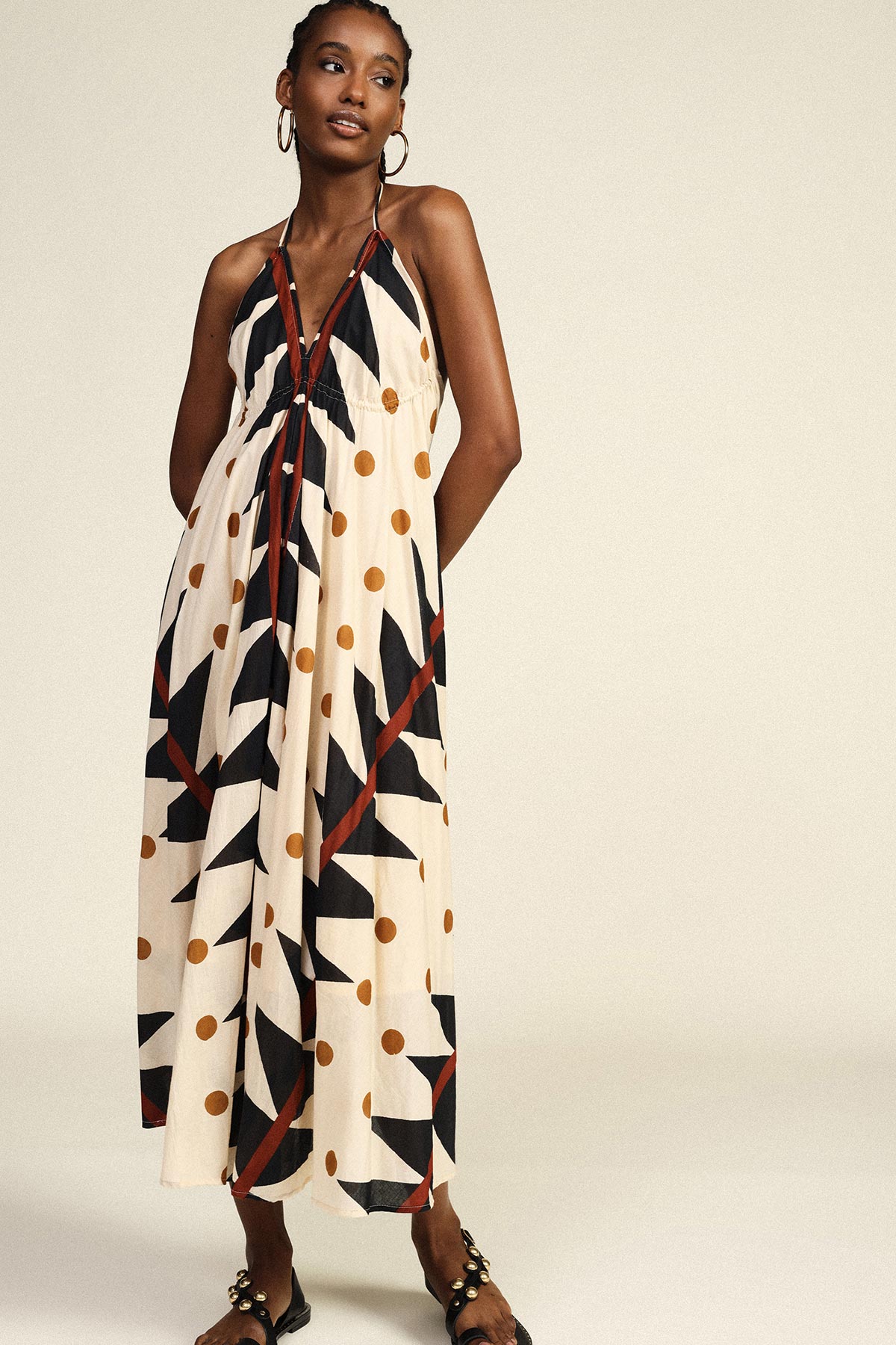 Woman in cream halter maxi dress with black geometric shapes and mustard polka dots, hoop earrings and black sandals, studio shot