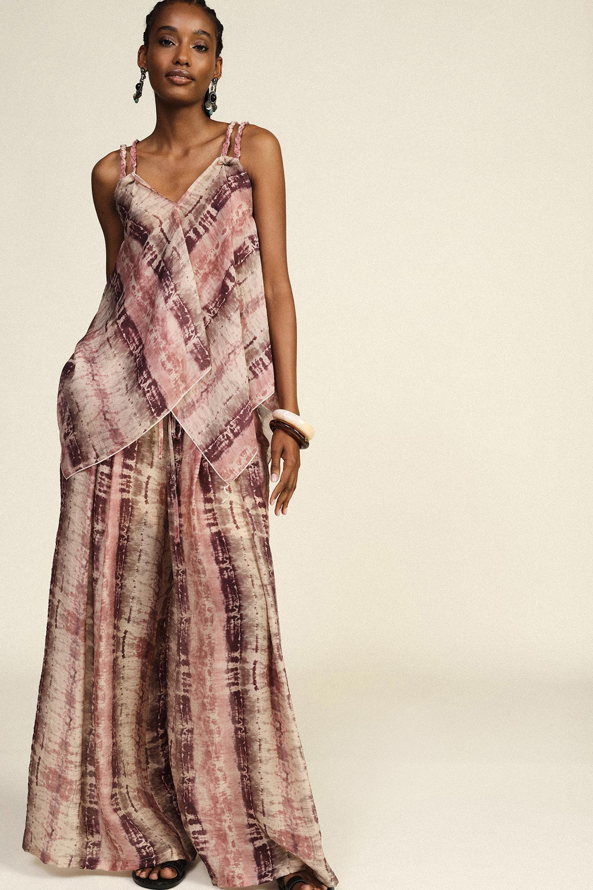 Woman wearing pink and mauve tie-dye layered cami top with braided straps and matching wide-leg trousers, accessorised with bangles and drop earrings