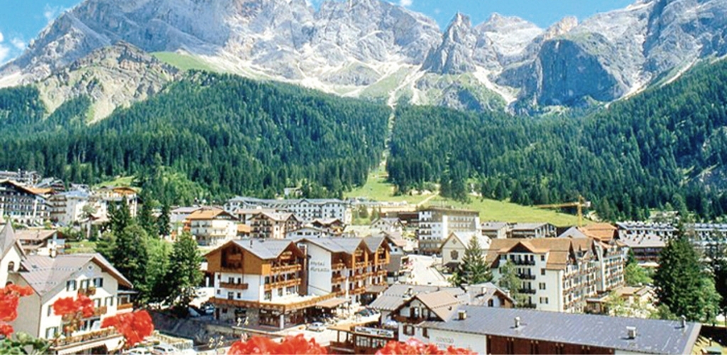 Alpine village of chalet-style houses beneath forested foothills and jagged rocky peaks, red flowers in foreground