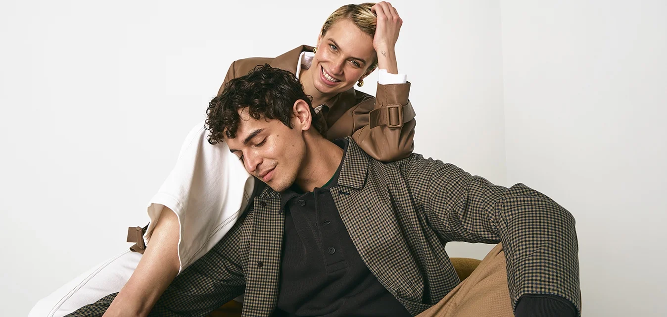 Due modelli sono seduti su una sedia marrone. Uno indossa una camicia a quadri, una polo nera e pantaloni beige; l'altro ha un cappotto marrone sopra un vestito bianco.