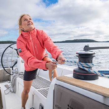 Person in a coral jacket trimming a sail on a sailboat winch with calm sea and cloudy sky in the background