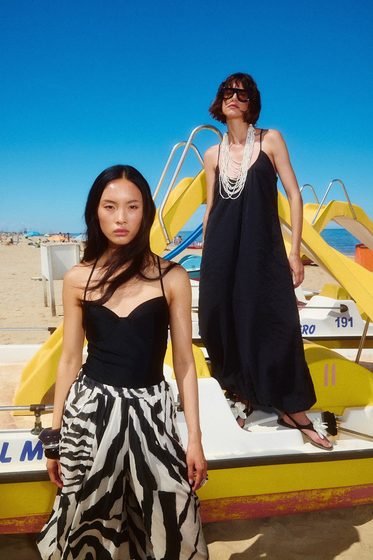 Two women in black summer dresses posing on a yellow pedal boat at a sunny beach with clear blue sky