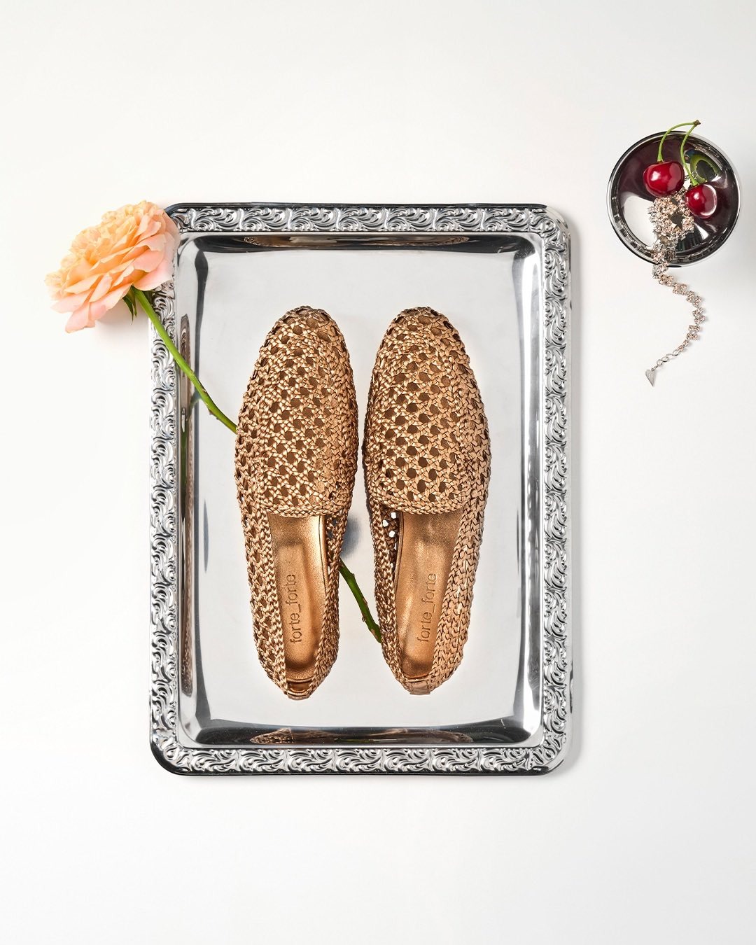 Woven bronze slip-on shoes on engraved silver tray with a peach rose stem and a small bowl of cherries and jewellery