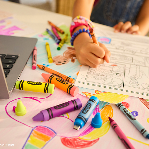 Primo piano di un bambino che colora con i pastelli Crayola, alcuni dei quali hanno lo stile di chiavette USB, su un tavolo con disegni e un laptop.