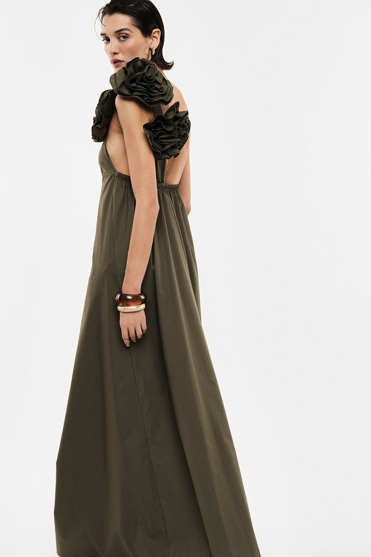 Woman wearing an olive-green sleeveless maxi dress with oversized ruffled shoulder details and open back, wearing wooden bangles