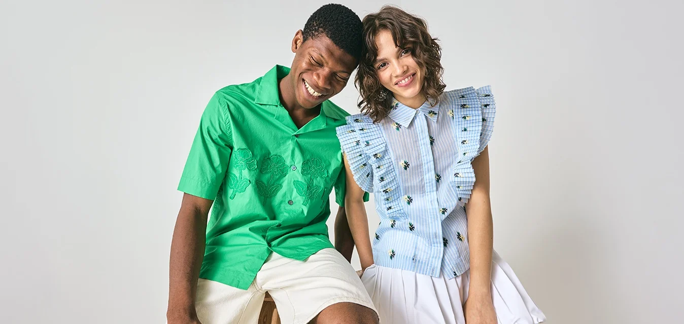 Giovane uomo in camicia verde e pantaloni beige e giovane donna in blusa blu con volant e gonna bianca siedono vicini, sorridendo davanti a uno sfondo semplice.