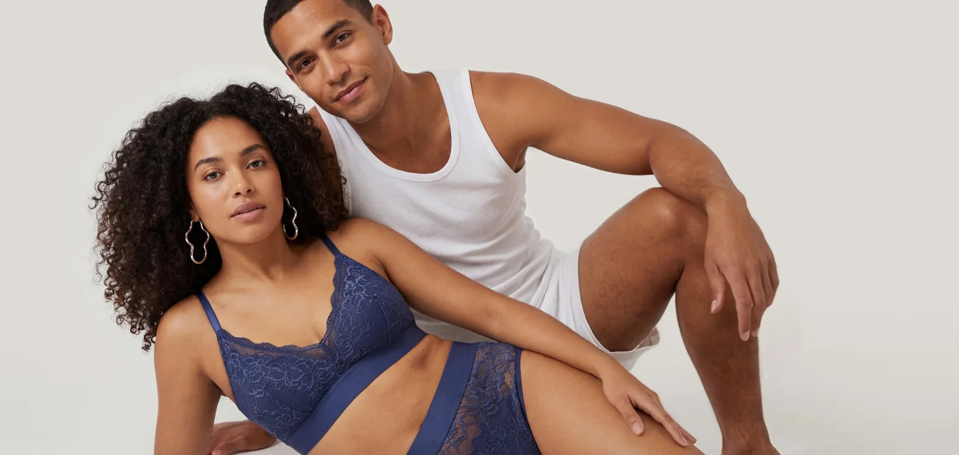 Donna in lingerie blu di pizzo che si rilassa mentre un uomo con canottiera bianca e pantaloncini è seduto dietro di lei su uno sfondo chiaro e semplice.
