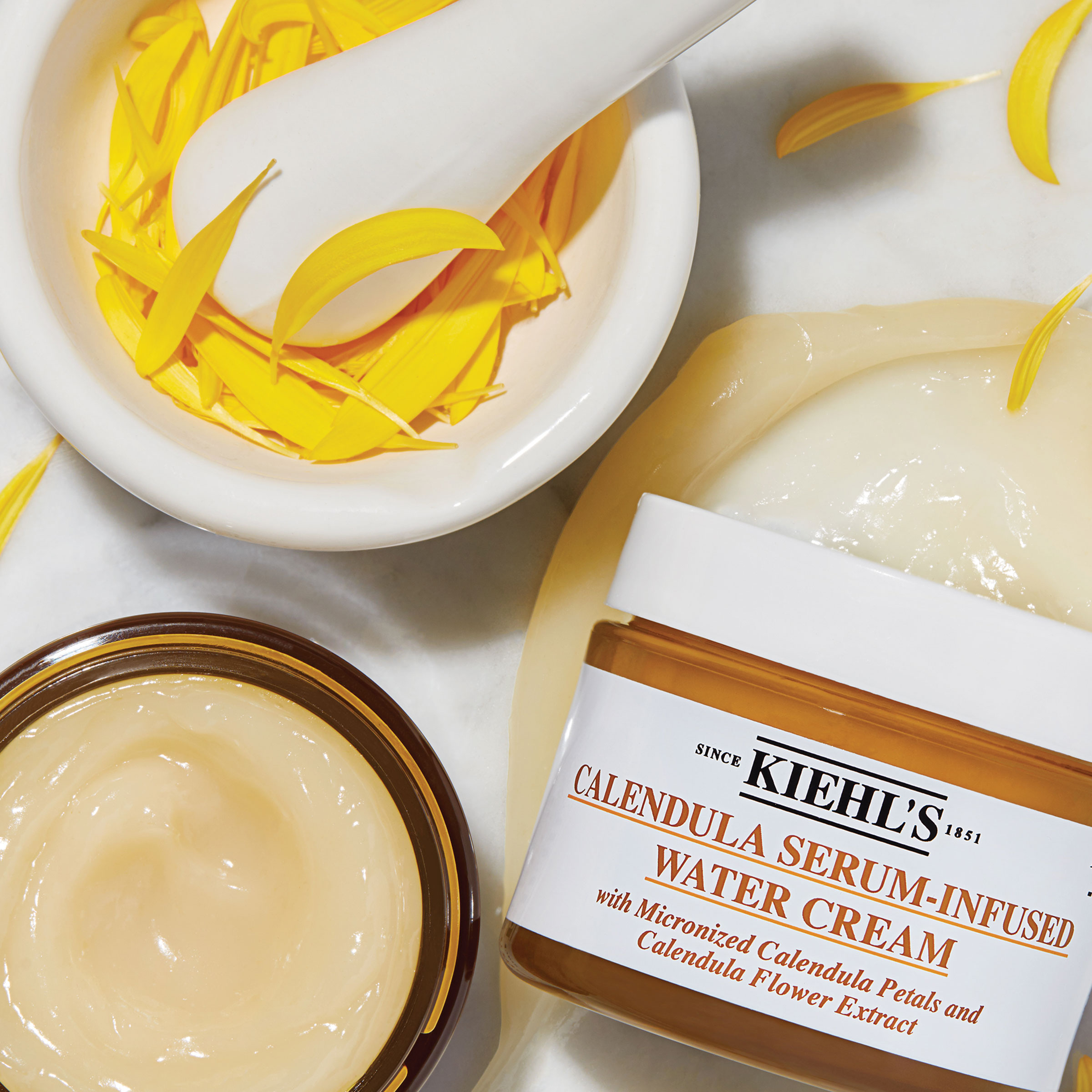 Flat lay con petali di Calendula gialli, mortaio bianco e due vasetti aperti di Calendula Serum-Infused Water Cream Kiehl's.