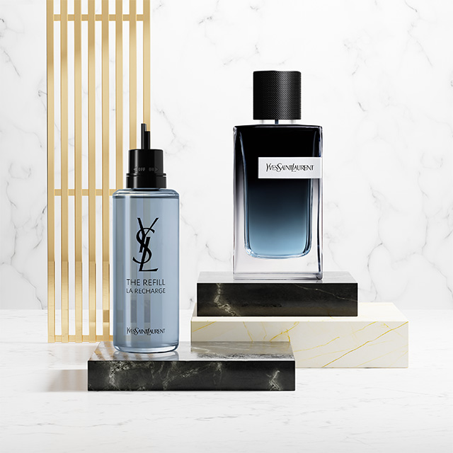 Un flacone di profumo blu YSL Y e la sua ricarica azzurra esposti su piedistalli in marmo e pietra nera.