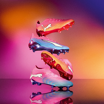 Vibrant stacked Puma football boots in pink, blue and gold on a reflective surface with a warm gradient background