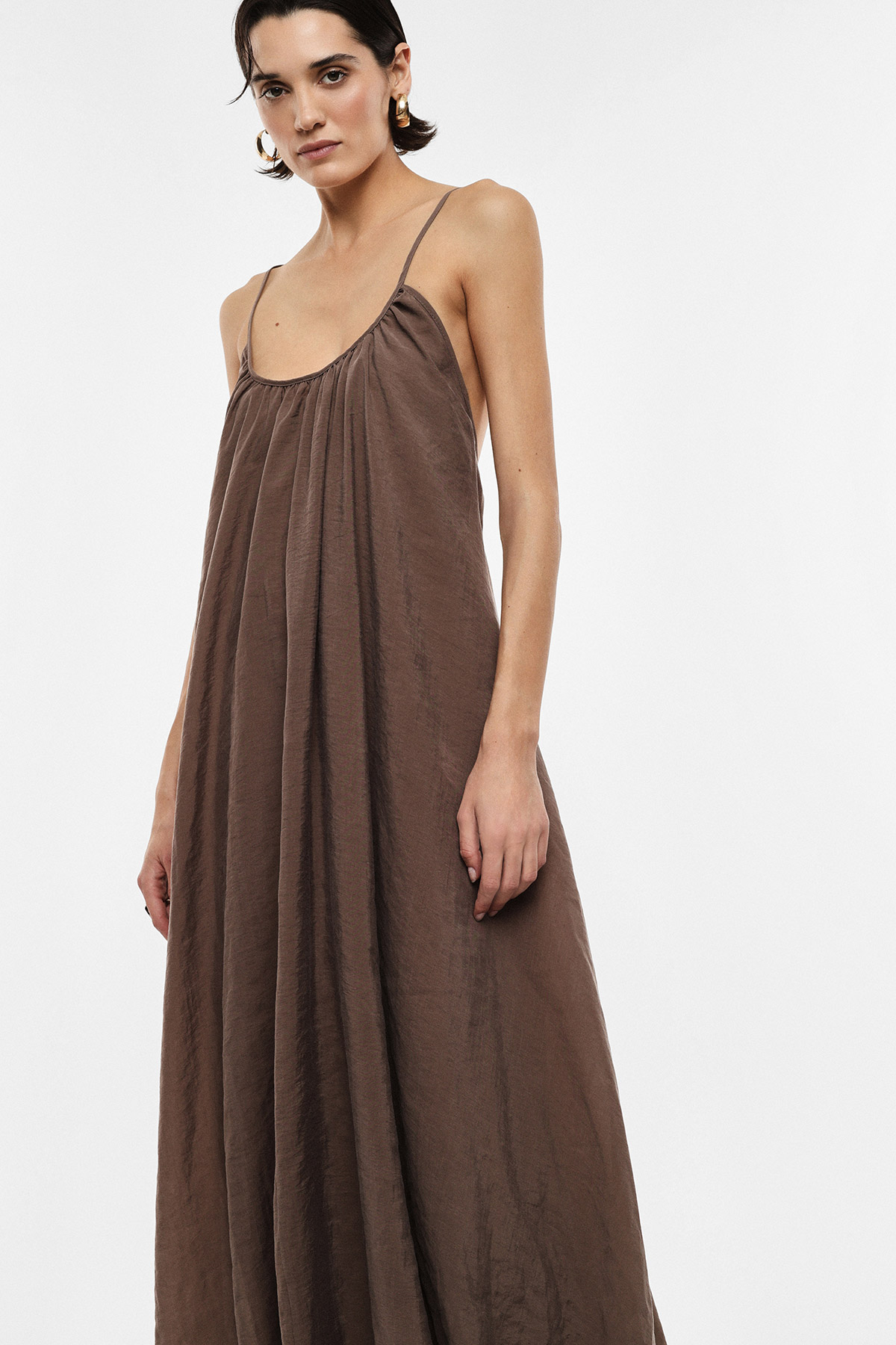 Woman modelling brown spaghetti-strap maxi dress with gathered neckline and gold hoop earrings against a white background