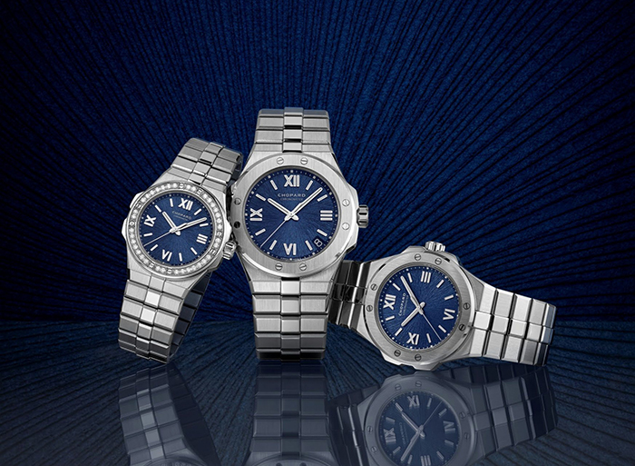 Three stainless-steel watches with blue sunburst dials, Roman numerals and metal bracelets on a reflective surface, radial blue background