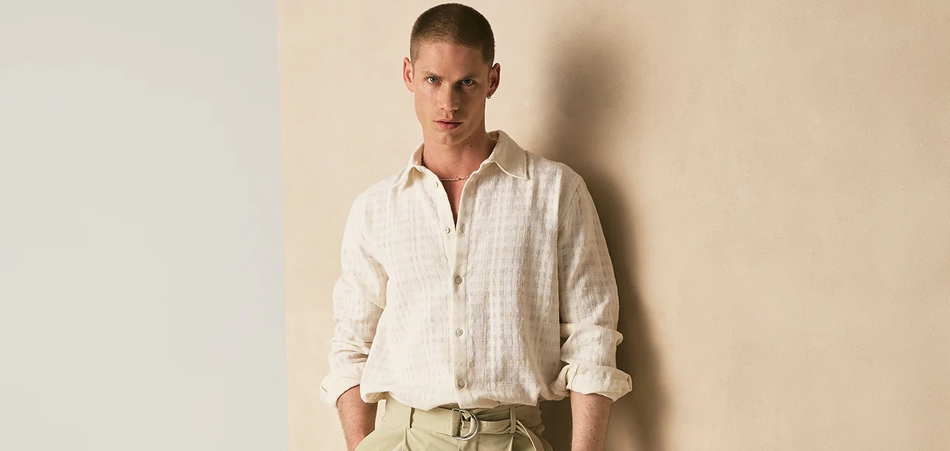Uomo con pantaloni beige a gamba larga e camicia bianca strutturata, in piedi con le mani nelle tasche davanti a un muro bicolore beige e bianco sporco.