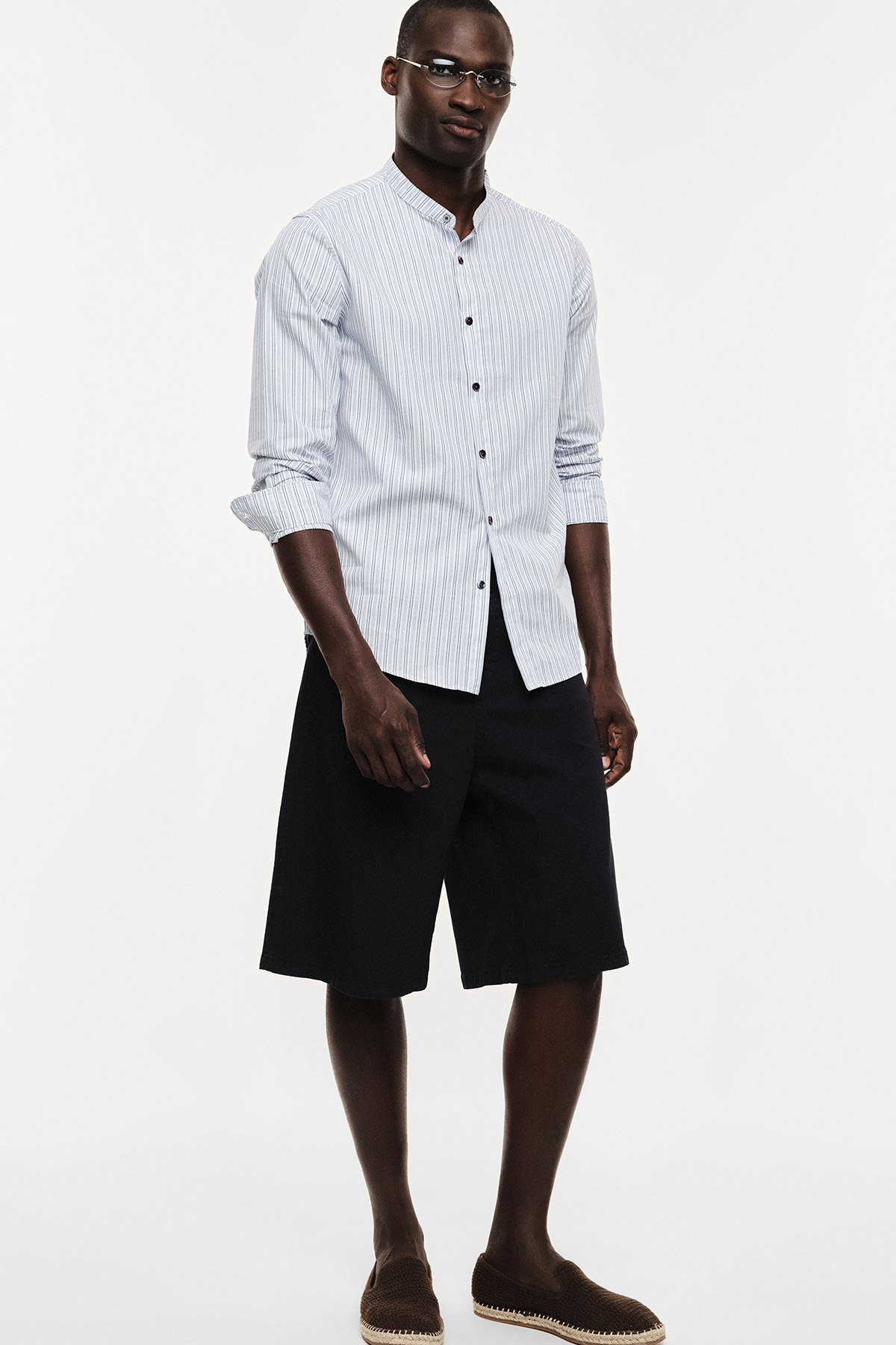 Male model wearing white striped band-collar shirt, black knee-length shorts, brown espadrilles and glasses